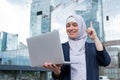 Business woman in hijab and suit is using a laptop and holding her index finger up outdoors. Royalty Free Stock Photo