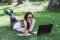 Business woman, lies in a summer grass park, using a laptop smiles and holds out a glass with coffee. Remote work during Royalty Free Stock Photo