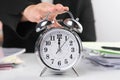 Business woman`s hand reaches out to touch alarm clock to stop time while sitting at her office desk. Selective focus of alarm Royalty Free Stock Photo