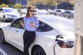 Business woman standing on city parking near electric car, charging automobile battery from small city station, drinking coffee a
