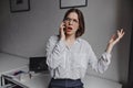 Business woman in stylish blouse emotionally talking on phone. Shot of girl with glasses on background of white table Royalty Free Stock Photo