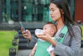 Business woman using telephone with carrying her infant and feeding her child`s by milk bottle. Busy mother hurrying in the