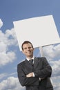 Businessman with arms crossed standing by blank sign against cloudy sky Royalty Free Stock Photo