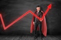 Businessman in black suit, red cape and red mask with glaring white eyes holding with one hand red graph arrow pointing