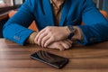 A businessman in a blue suit, jacket with a watch in his hands sits idly by next to a mobile phone or smartphone lying on a wooden Royalty Free Stock Photo