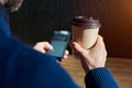 Businessman in blue sweater drinks coffee in a sunny cafe, close-up holds a disposable paper glass, using smartphone while having Royalty Free Stock Photo
