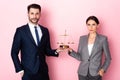 Businessman and businesswoman with hands in pockets holding golden scales on pink