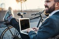 Businessman commuter with bicycle sitting on bench in city, using laptop. Copy space. Royalty Free Stock Photo