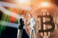 businessman figurines shaking hands in front of blurred stock market chart and bitcoin Royalty Free Stock Photo