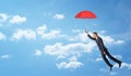 A businessman flying in the clouded sky and holding a red open umbrella.