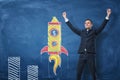 Businessman with hands raised in victory and rocket drawn in chalk on blue blackboard background