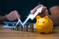 Businessman holding a coin in a piggy bank with graphs and arrows showing charts on his desk. Royalty Free Stock Photo