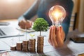 businessman holding lightbulb with turbine and tree grow on coins. concept saving energy and finance accounting