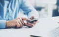 Businessman holding smartphone in hands and touching screen. Generic design laptop on the table. Blurred background