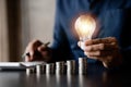 Businessman holds a glowing light bulb on top of the highest pile of coins, Placing coins in a row from low to high is comparable Royalty Free Stock Photo