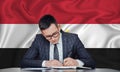 A businessman in a jacket and glasses sits at a table signs a contract against the background of a flag Egypt