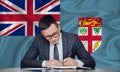 A businessman in a jacket and glasses sits at a table signs a contract against the background of a flag Fiji