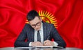 A businessman in a jacket and glasses sits at a table signs a contract against the background of a flag Kyrgyzstan