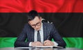 A businessman in a jacket and glasses sits at a table signs a contract against the background of a flag Libya