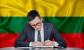 A businessman in a jacket and glasses sits at a table signs a contract against the background of a flag Lithuania, Vilnius,