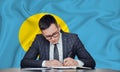 A businessman in a jacket and glasses sits at a table signs a contract against the background of a flag Palau