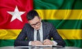 A businessman in a jacket and glasses sits at a table signs a contract against the background of a flag togo