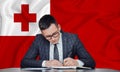 A businessman in a jacket and glasses sits at a table signs a contract against the background of a flag tonga, Nuku`alofa