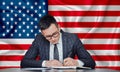 A businessman in a jacket and glasses sits at a table signs a contract against the background of a flag US
