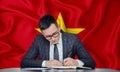 A businessman in a jacket and glasses sits at a table signs a contract against the background of a flag Vietnam