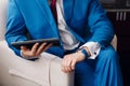 Businessman keep a digital tablet in hand whilst sitting on a sofa in a blue suit. on hand expensive mechanical watch