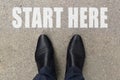 Businessman is looking down at his feet on a concrete floor with START HERE letters painted on the surface. Top view image of pers