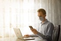 Businessman in medical mask during quarantine. A young man in a cafe works at a laptop by the window. Isolation, coronavirus, work Royalty Free Stock Photo