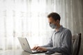 Businessman in medical mask during quarantine. A young man in a cafe works at a laptop by the window. Isolation, coronavirus, work Royalty Free Stock Photo