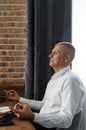 A businessman meditates while working on a laptop, sitting with his eyes closed at his workplace in a home interior.