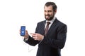 Cheerful bearded businessman listening music with earphones and pointing at smartphone with facebook website