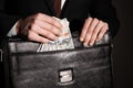 Businessman putting dollar banknotes into briefcase on dark background, closeup. Corruption concept