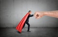 A businessman in a red cape and a mask pushing at a giant human fist on concrete background.