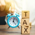 Businessman`s hand holding a clock near the wooden blocks with the word Tax. Time to pay taxes. The concept of annual taxation.