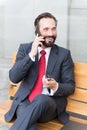 Businessman sitting on bench using his cell phone. Smiling bearded businessman sitting on bench and talking on the phone