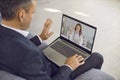 Businessman sitting on office couch, waving hand at laptop and greeting online doctor