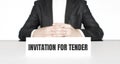 Businessman sitting at the table and signboard with text INVITATION FOR TENDER . Business concept