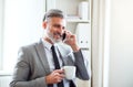 Businessman with smartphone and coffee standing in an office, making a phone call.