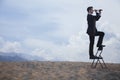 Businessman standing on a chair and looking through a telescope in the middle of the desert