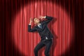 Businessman standing in spotlight near red stage curtain, leaning aside, looking up and protecting head with hands from Royalty Free Stock Photo