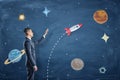 A businessman stands near a blackboard with drawings of planets and points to the rocket.