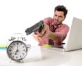 Businessman in stress at office computer desk pointing hand gun to alarm clock in out of time and project deadline expiring