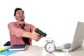 Businessman in stress at office computer desk pointing hand gun to alarm clock in out of time and project deadline expiring