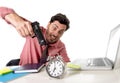 Businessman in stress at office computer desk pointing hand gun to alarm clock in out of time and project deadline expiring