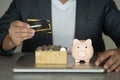 Businessman in suit holding credit card and pink piggy bank with stock of coin in box place on notebook