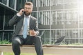 Businessman in suit and tie is sitting outside on bench, drinking coffee and talking on his cell phone. Nearby is laptop Royalty Free Stock Photo